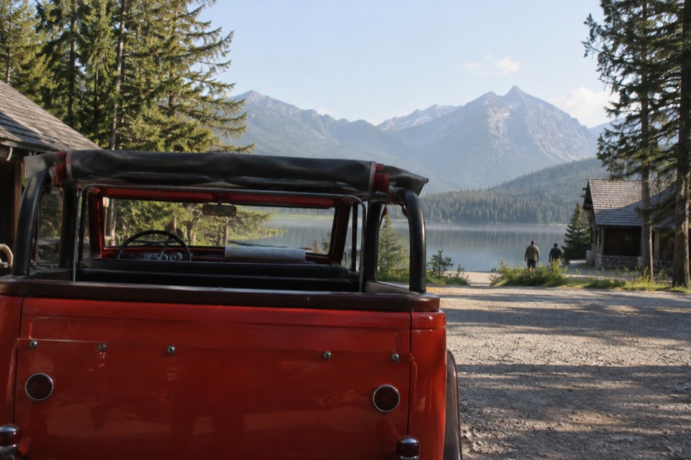 A red vintage tour bus parked beside a mountain lake with tall peaks behind it