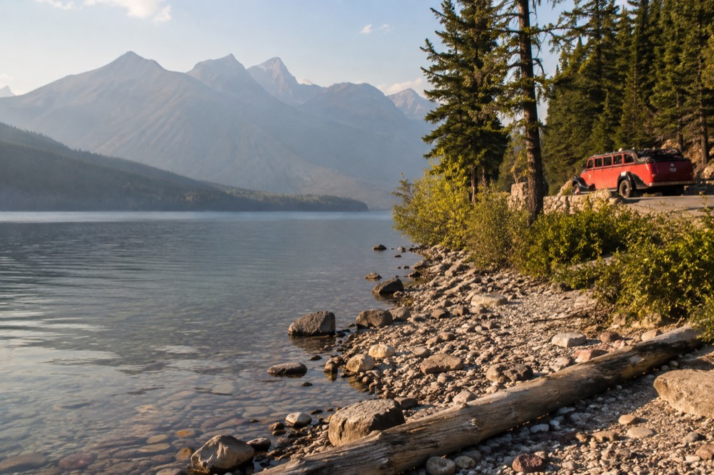 A red vintage tour bus parked beside a lake with mountains in the distance