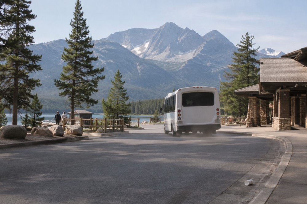 A shuttle bus pulling away from a mountain road pullout with tall peaks behind it
