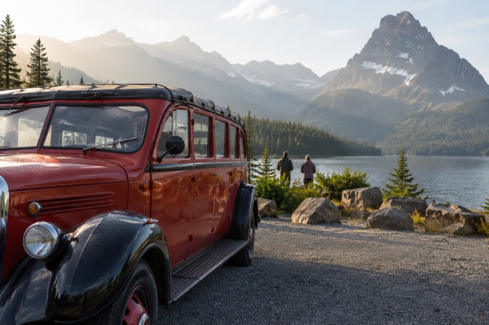 A vintage red tour bus parked beside a mountain lake with forested peaks behind it