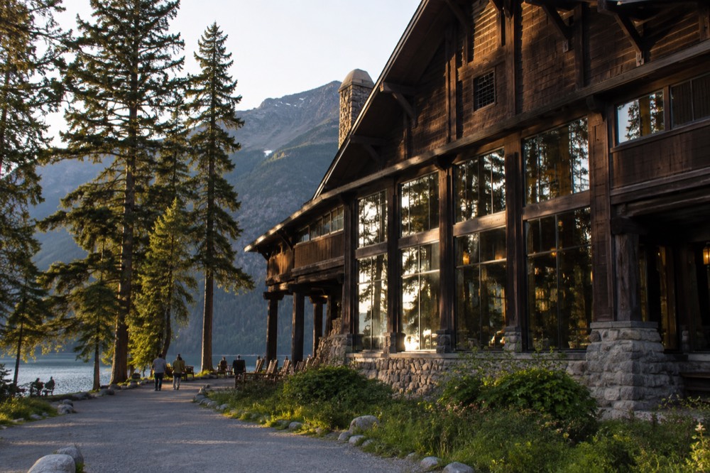 A wooden historic lodge with a stone fireplace and tall windows beside a forested lake