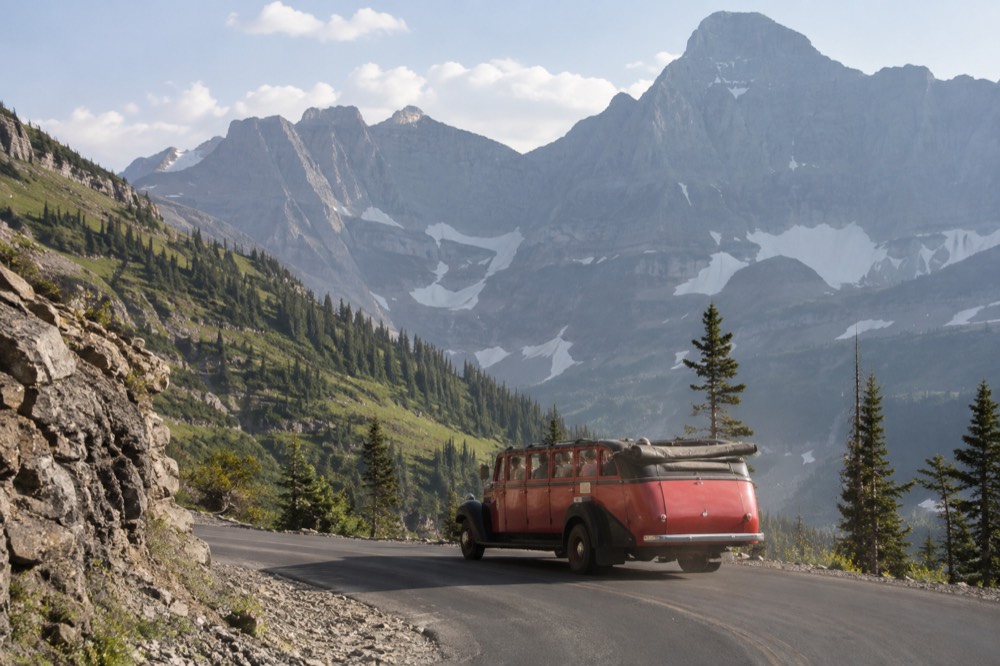 A red open-top vintage bus rounding a curve on a mountain road with peaks beyond