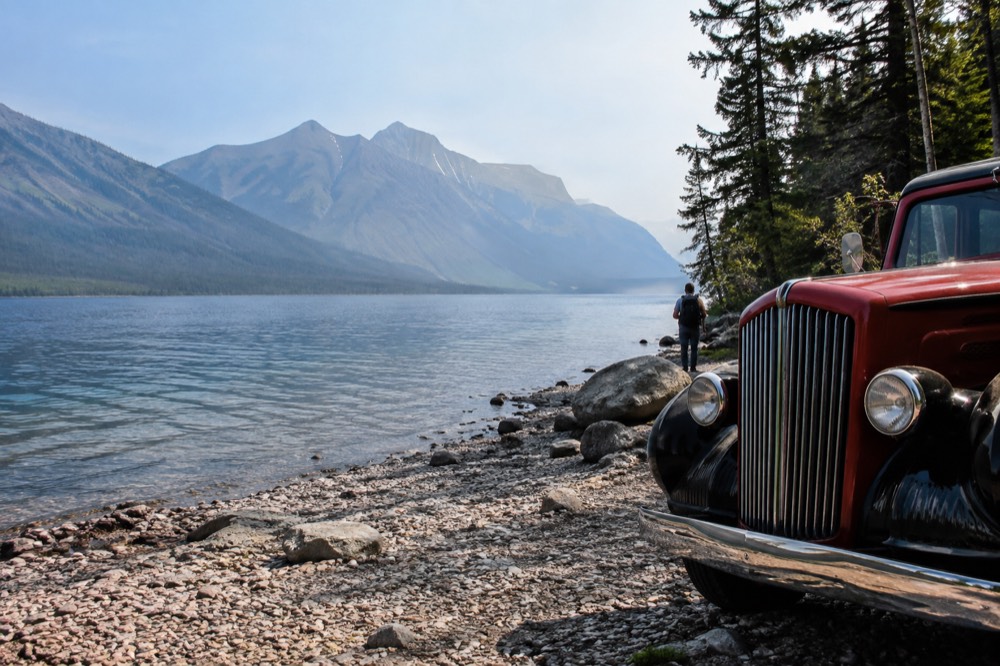 A vintage style tour vehicle parked beside a lake with mountains behind it