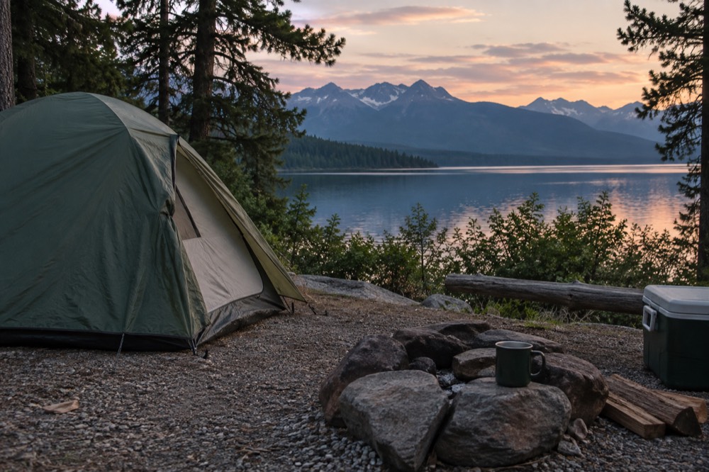 A campsite beside a lake with a tent and mountain peaks reflected in still water