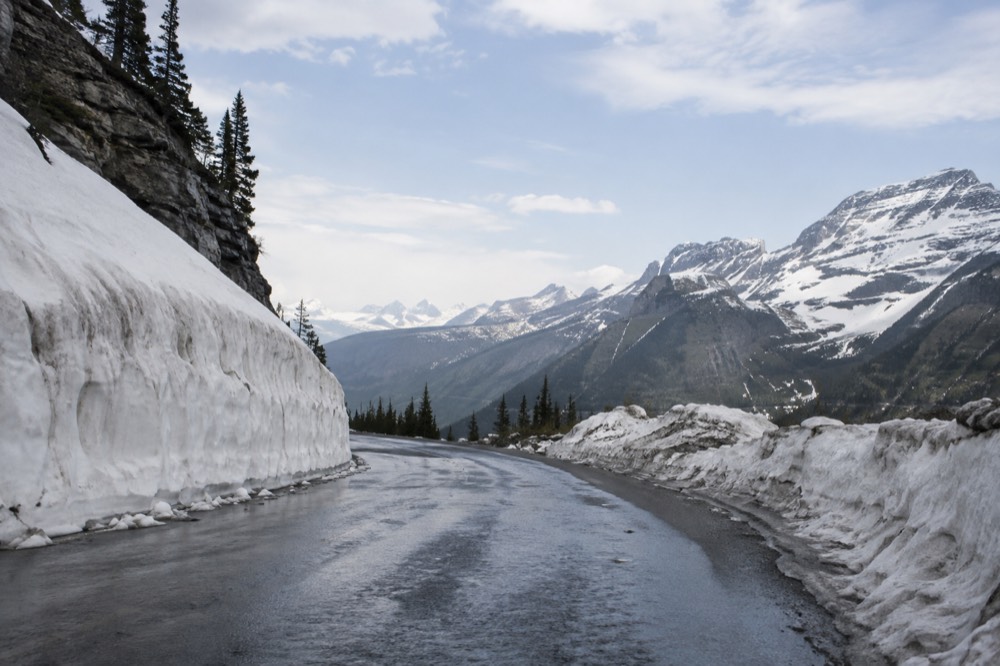 Snowbanks lining both sides of a high mountain road with a valley visible below