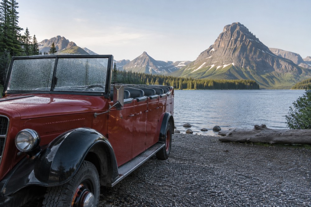 A vintage open-top tour bus parked beside a lake with mountains behind