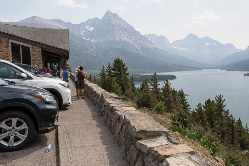 A busy overlook with vehicles parked along a stone wall and mountains beyond