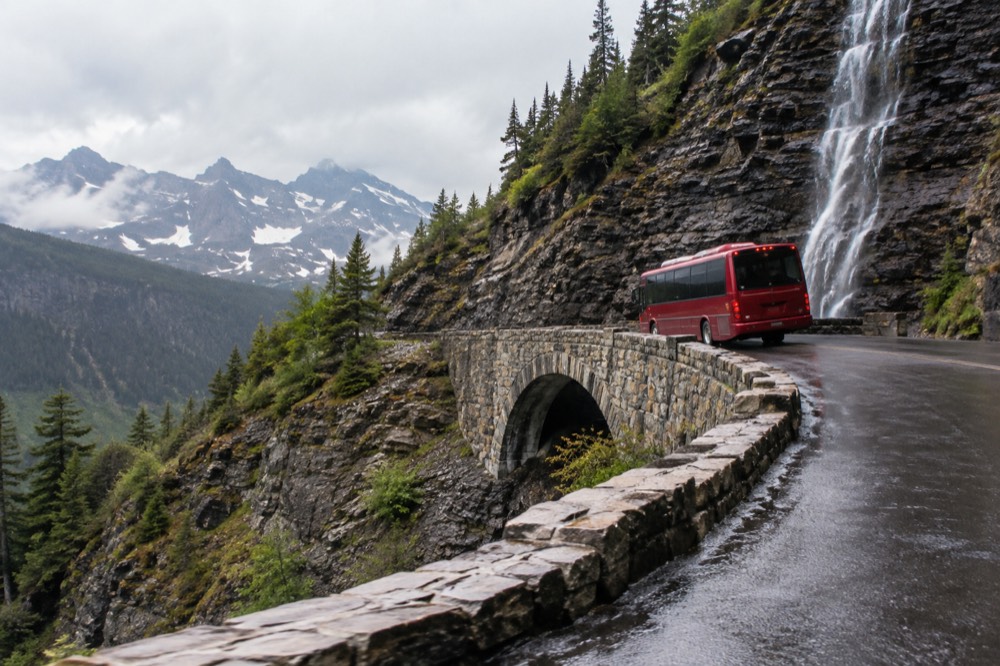 A red bus crossing a stone bridge on a mountain road with a waterfall in the background