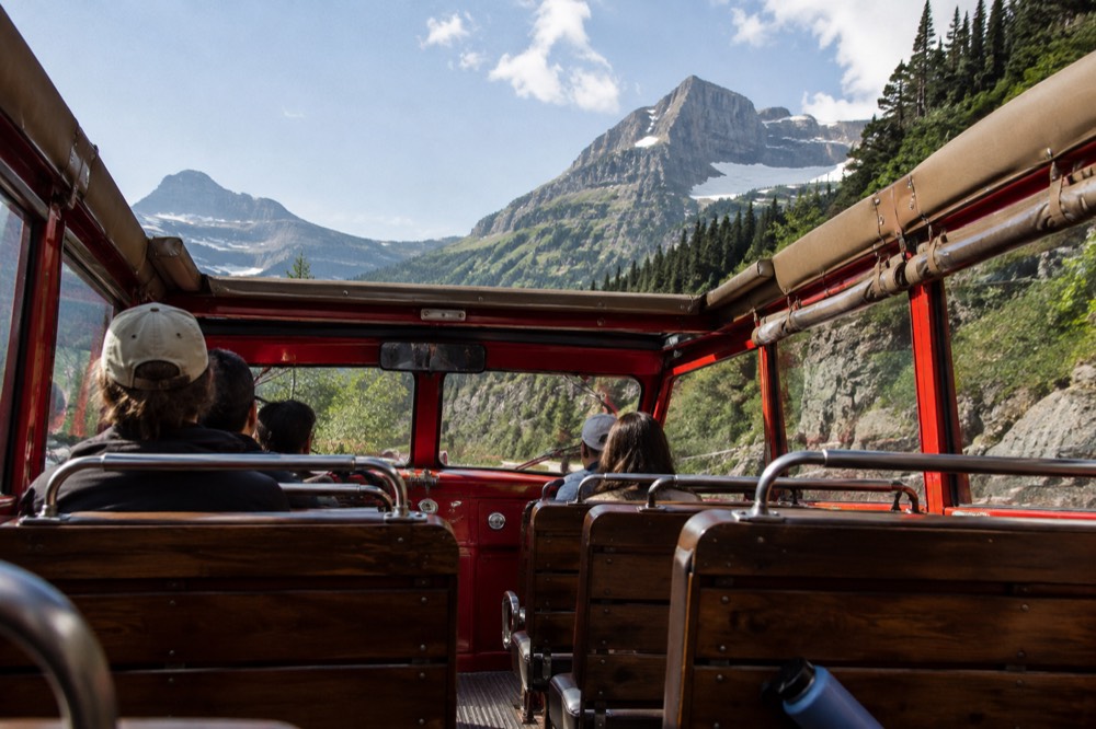 A red bus interior with wooden bench seats and an open canvas roof showing mountains