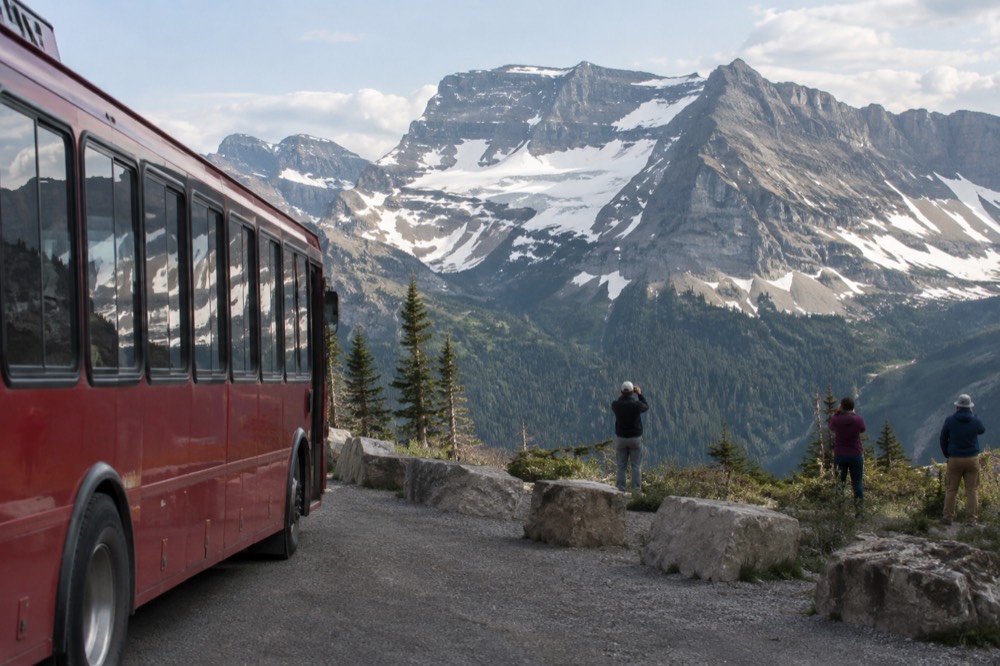 A red bus stopped at a pullout while passengers photograph a mountain vista